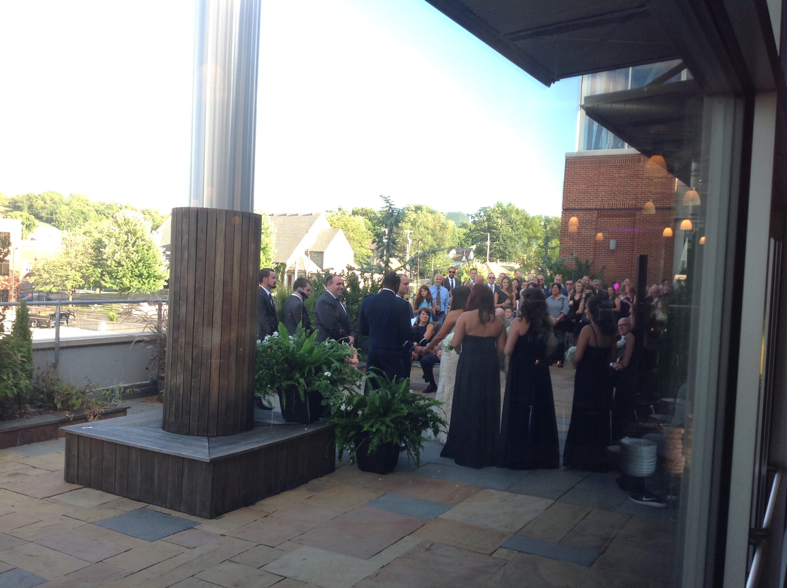A group of people, some in formal attire, gather outdoors on a patio for a wedding ceremony. The setting includes plants, seating, and buildings in the background on a sunny day.