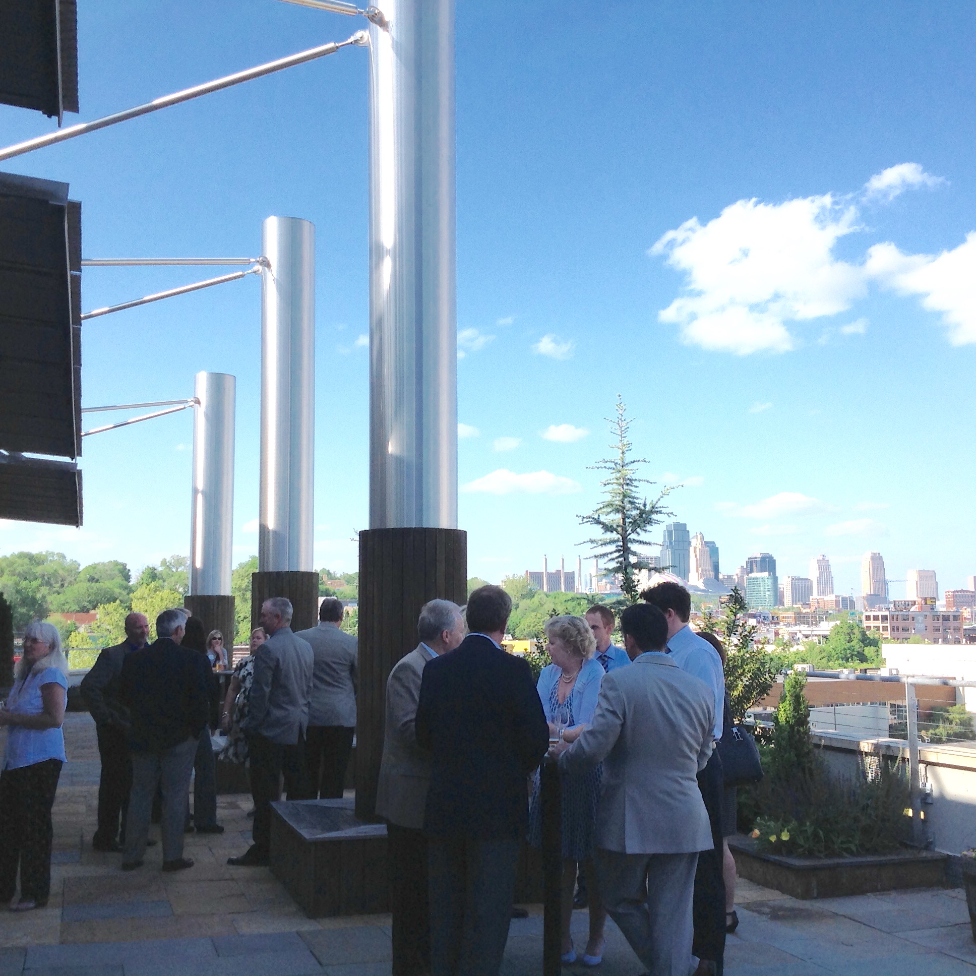 A group of people in business attire gather and converse on a terrace with modern metal columns, overlooking a city skyline under a bright blue sky with scattered clouds.