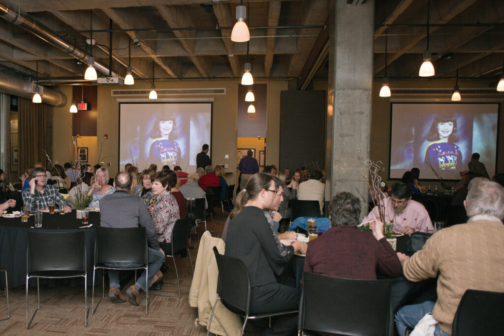 People sit at round tables in a large, industrial-style room, eating and talking. Two screens display an image of a young girl in a sweater with bangs. The atmosphere appears to be a social event or gathering.