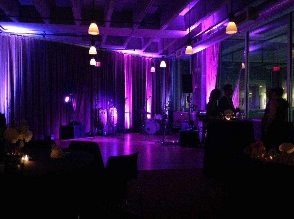A dimly lit event space with purple lighting, musical instruments set up on a stage, and silhouettes of people standing and conversing near round tables decorated with flowers and candles.