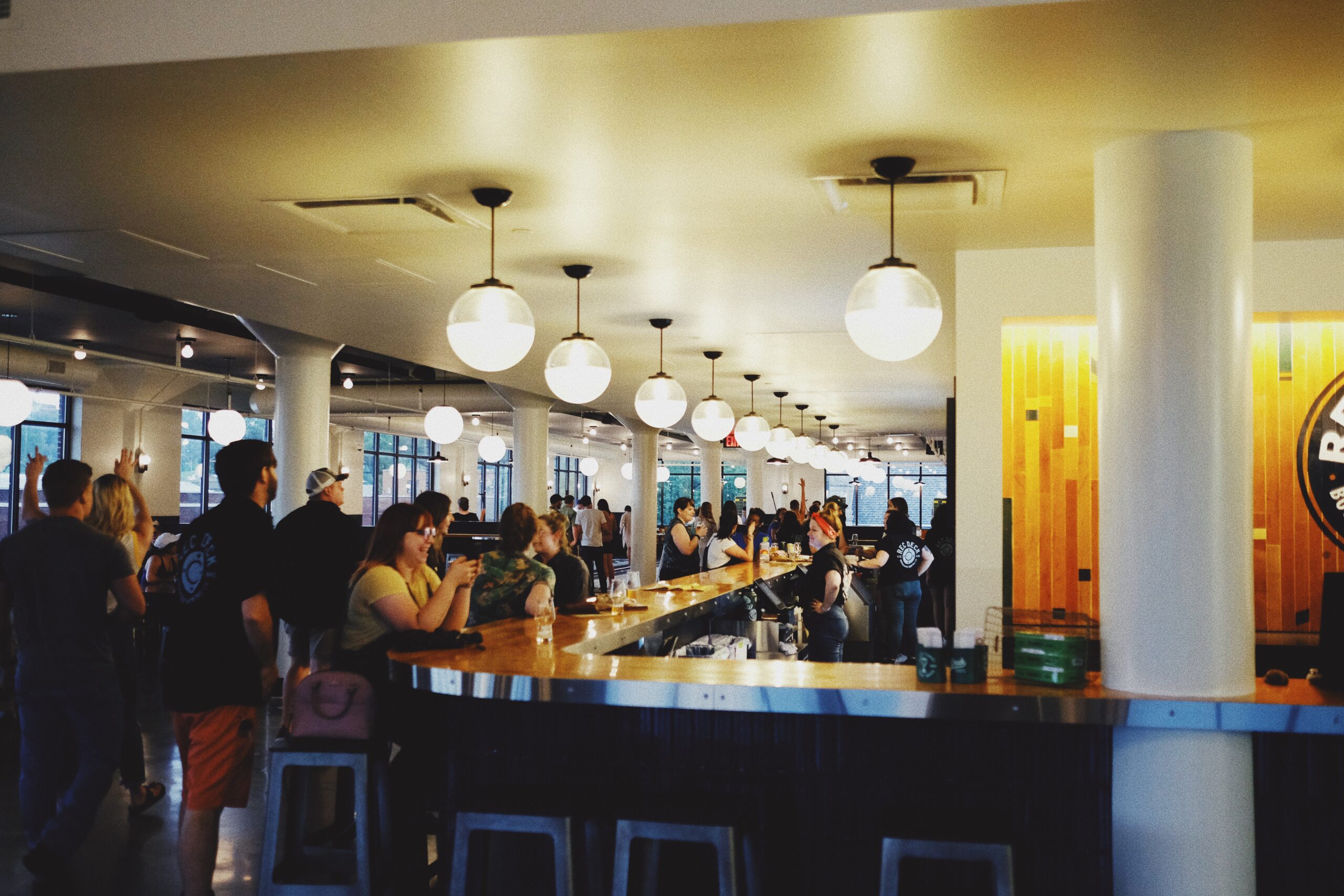 A busy, modern bar with groups of people socializing around a long wooden counter; round hanging lights and large windows brighten the space.