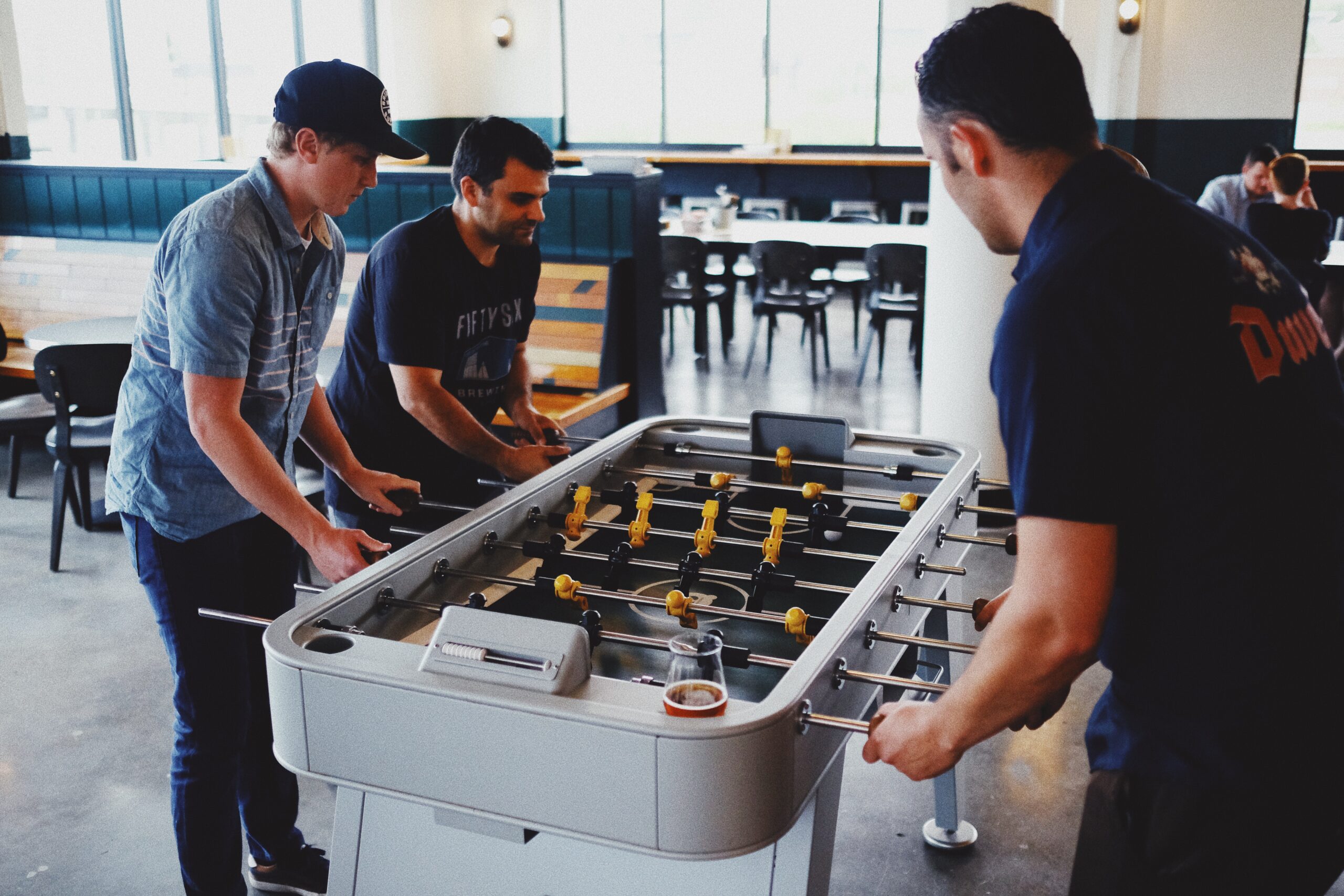 Three men are playing a game of foosball in a bright, modern indoor space with large windows. A glass of beer sits on the foosball table, and empty chairs and tables are visible in the background.