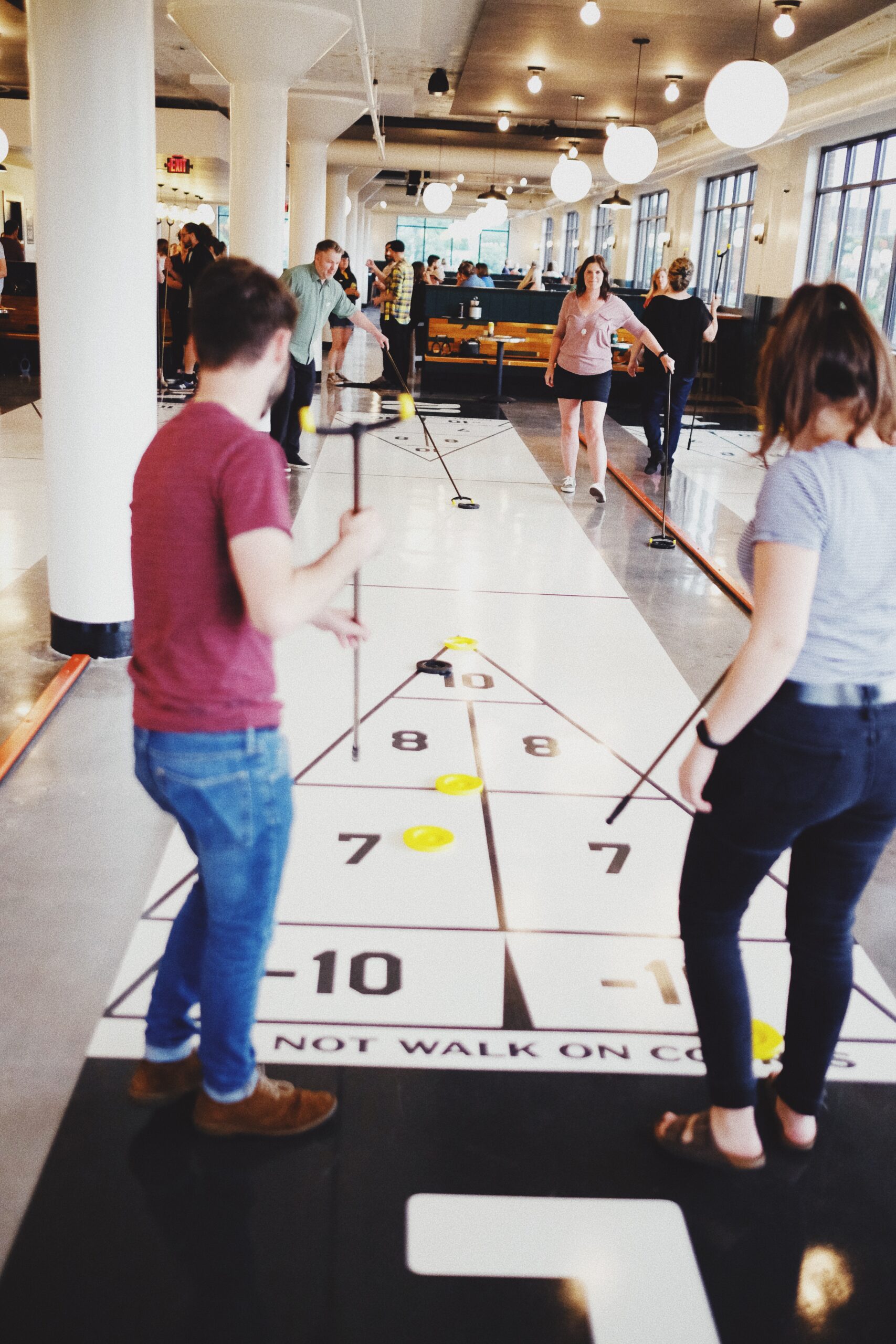 People play shuffleboard indoors, using long cues to push yellow pucks on a white court. Several people watch or play, and the space has large windows, white pillars, and round ceiling lights.