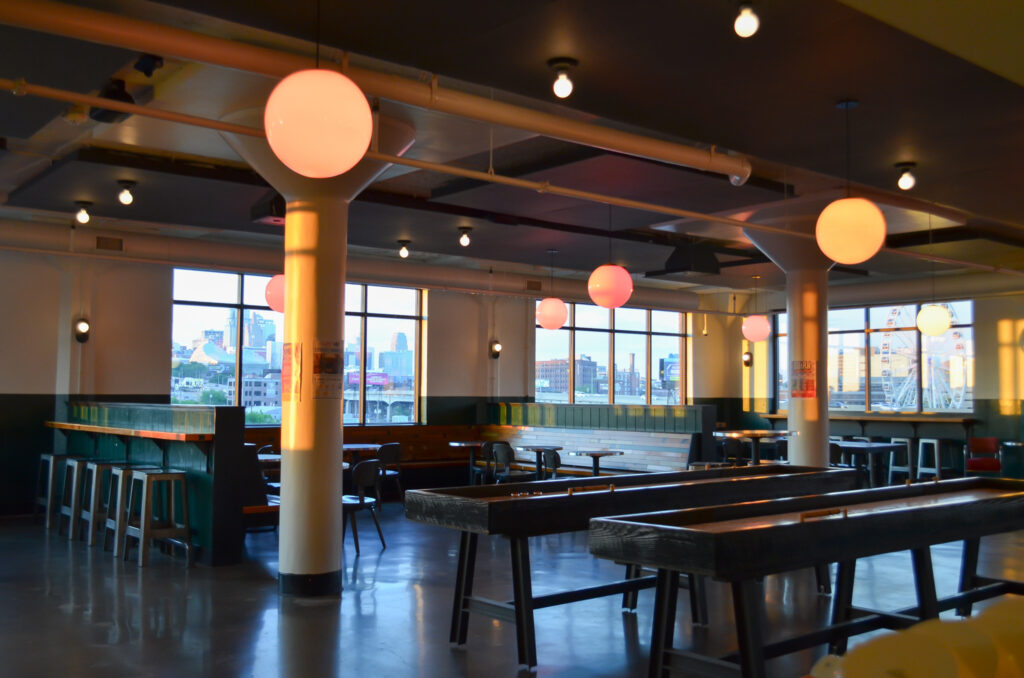 A spacious, modern dining area with large windows, high tables and stools, bench seating, and round pendant lights hanging from the ceiling, illuminated by natural light.