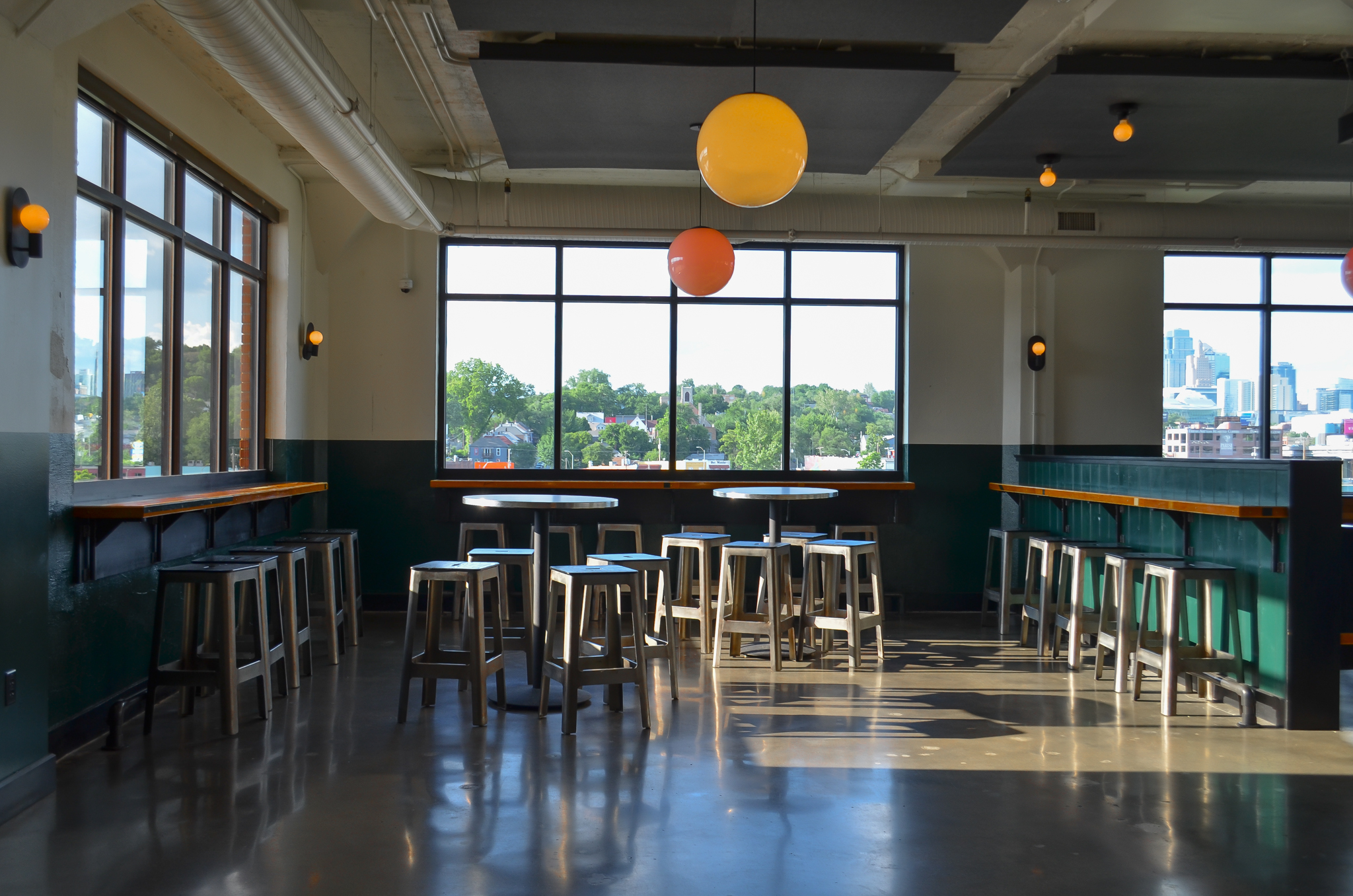 A bright, modern cafe with large windows, high tables, and stools. Three round pendant lights hang from the ceiling, and sunlight streams in, casting shadows on the polished floor. City buildings are visible outside the windows.