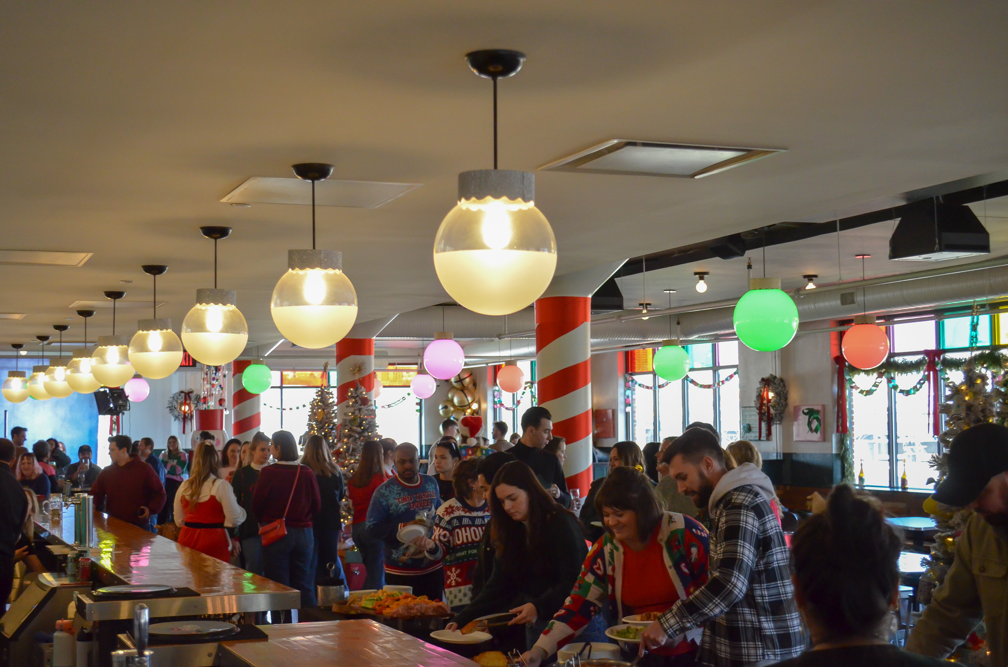A festive, crowded dining hall decorated with Christmas ornaments and candy cane pillars. People are serving themselves food at a buffet, and colorful lights hang from the ceiling.