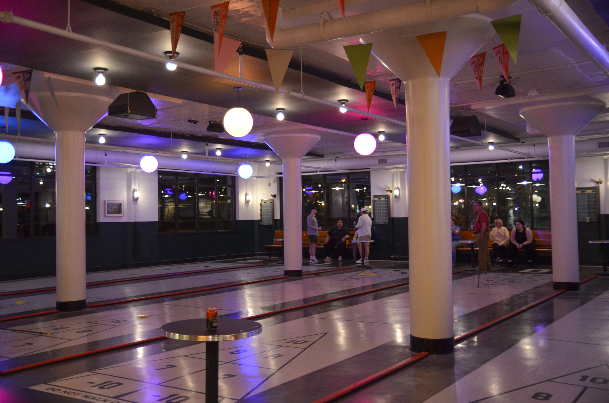 A spacious indoor shuffleboard venue with multiple lanes, round hanging lights, pillars, and festive pennant banners. People are gathered and seated at the back, chatting and watching the games.