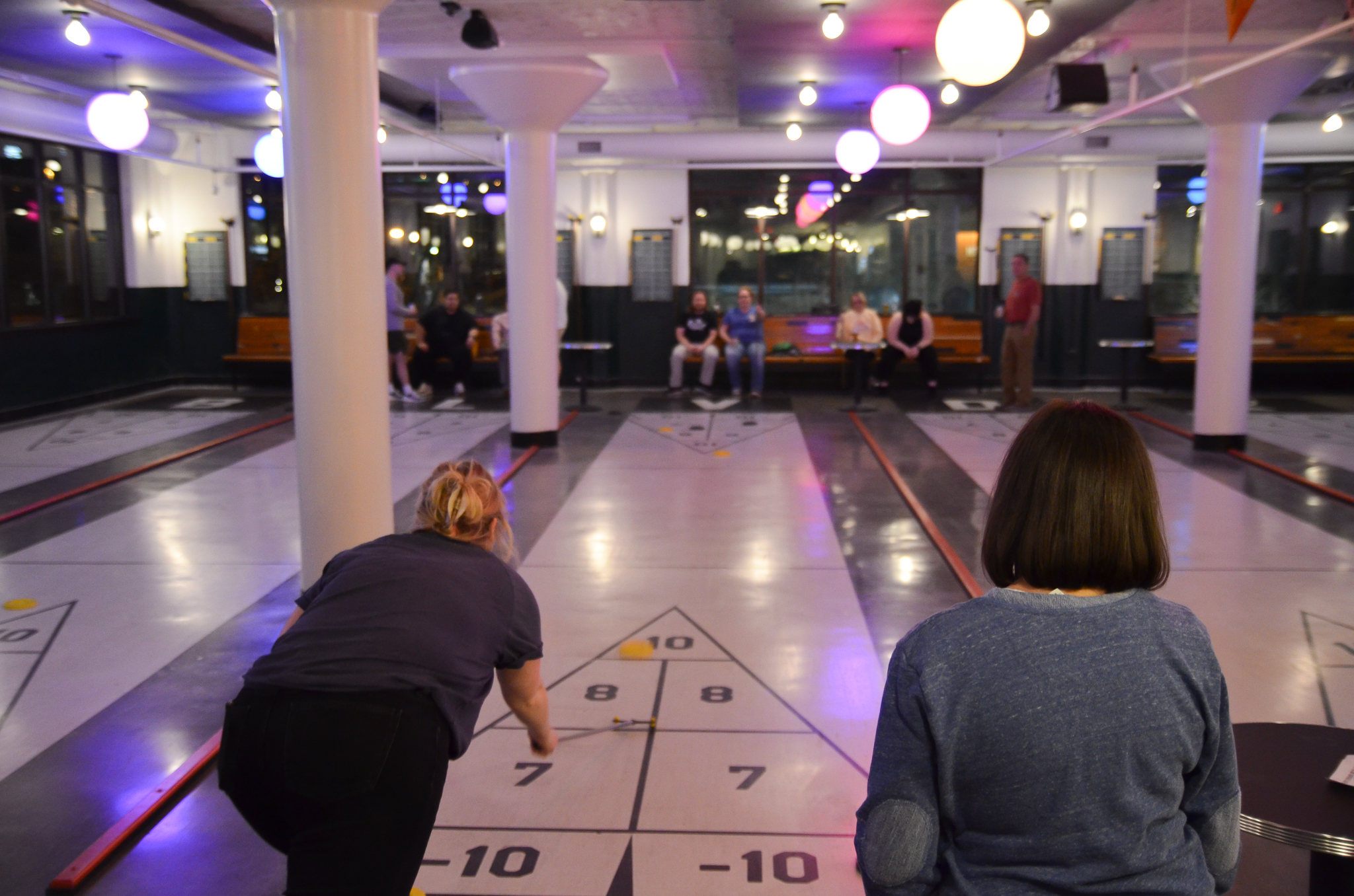 Two women play shuffleboard indoors, aiming discs toward a triangular scoring area as several people watch from benches in the background under hanging lights.
