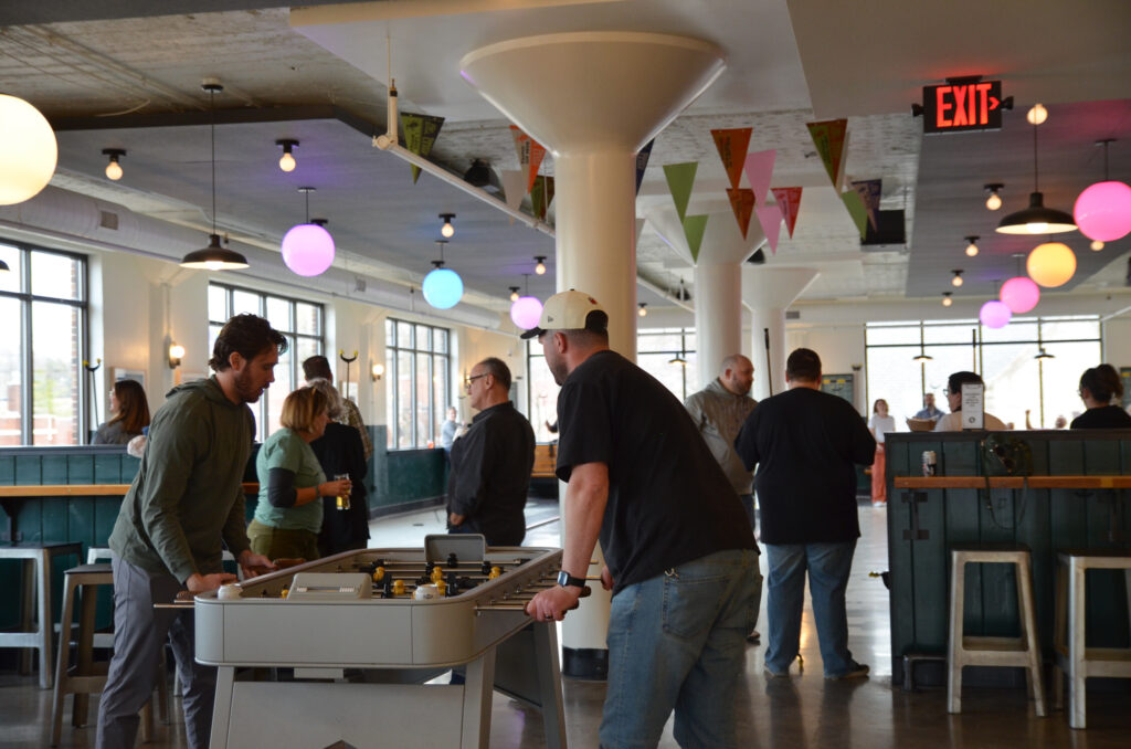 People gather in a bright, modern room with string lights and colorful flags. Two men play foosball in the foreground while others socialize in small groups in the background.
