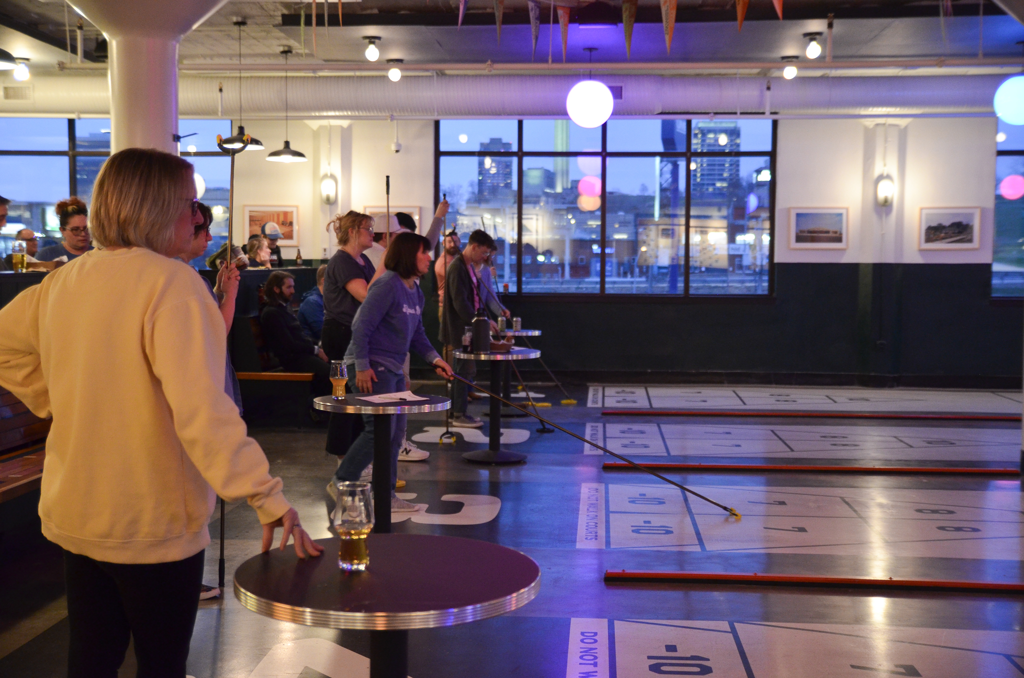 People play shuffleboard indoors at a lively venue. Several players focus on their turns while others watch. Drinks are on round tables, and large windows and wall art decorate the space. The lighting is warm and relaxed.