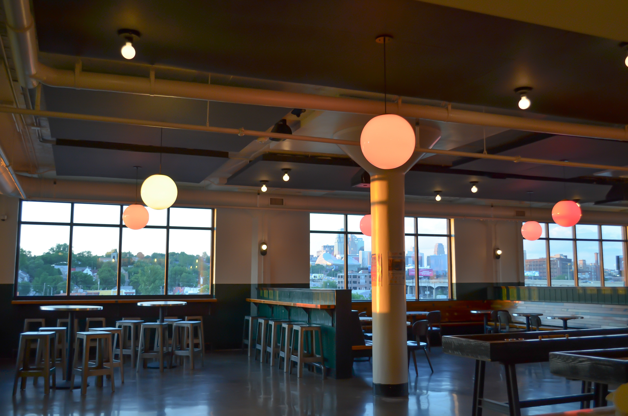 A modern, empty cafe with large windows, high-top tables, wooden stools, and round hanging lights. Sunlight streams in, showing a cityscape and trees outside. The interior has sleek, industrial-style decor.