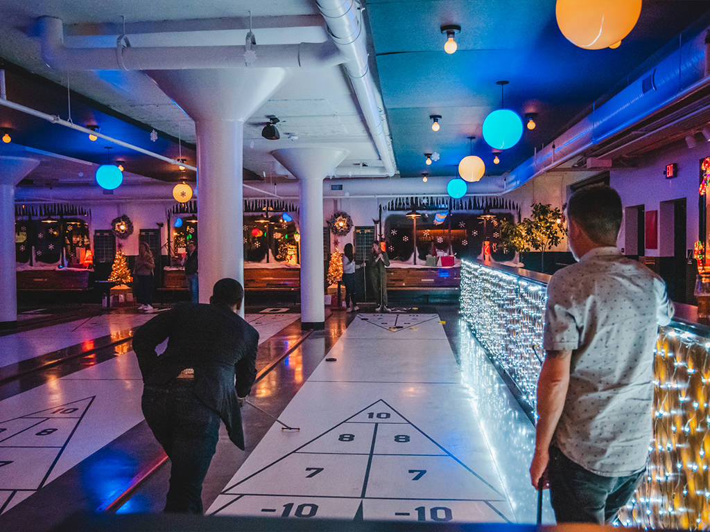 Two people play shuffleboard indoors under colorful lights, with festive wreaths and holiday decorations visible in the background. One person is taking a shot while another watches near a counter wrapped in string lights.