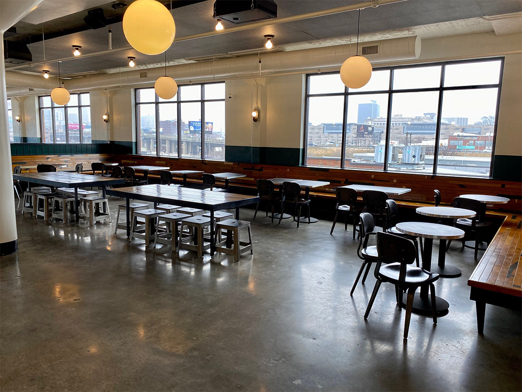 A spacious, modern cafeteria with large windows, wooden tables, black and metal chairs, and round ceiling lights. The floor is polished concrete, and the room is mostly empty, with a cityscape visible outside.