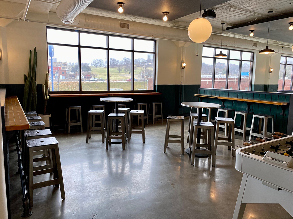 Modern cafe with high windows, metal stools, round and rectangular tables, a foosball table in the corner, and a large cactus by the wall. Bright natural light fills the space, showing an outdoor view with trees and buildings.