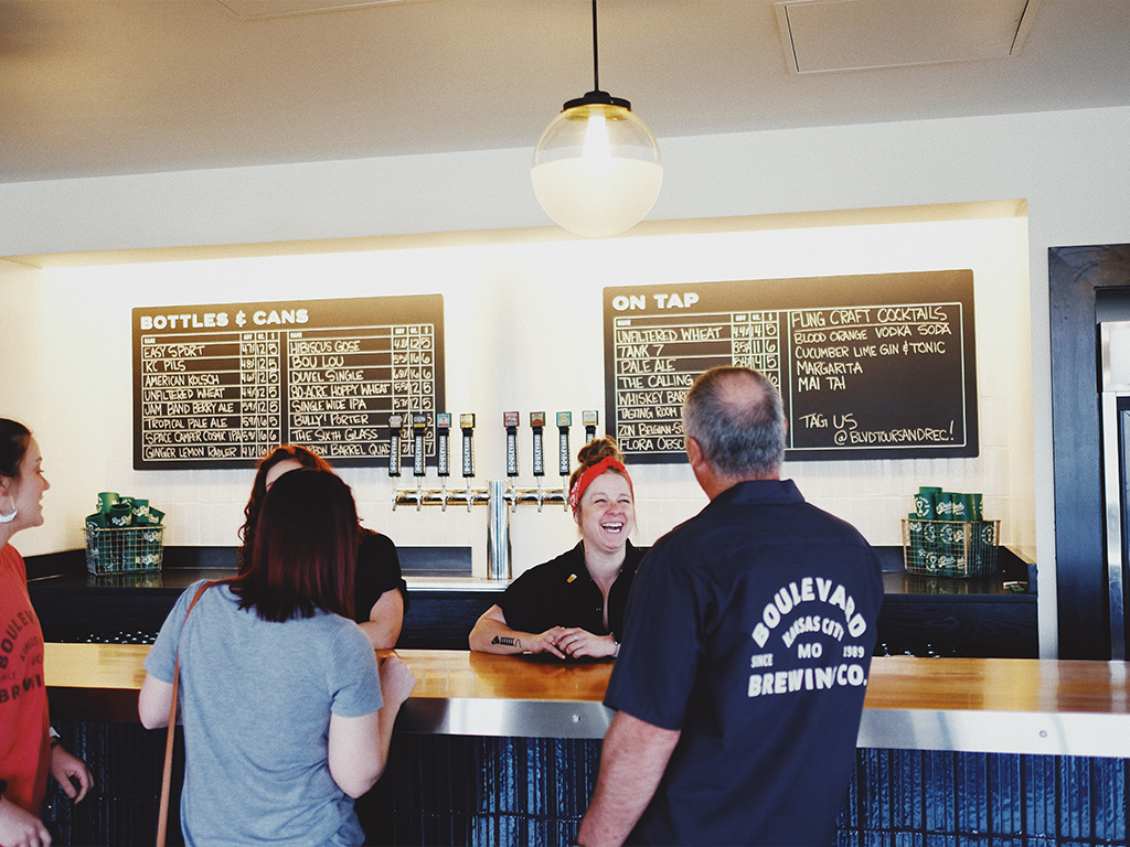 People are gathered at a bar counter, engaging in conversation. A smiling bartender stands behind the counter with drink menus displayed on chalkboards above. A row of beer taps is visible in the background.