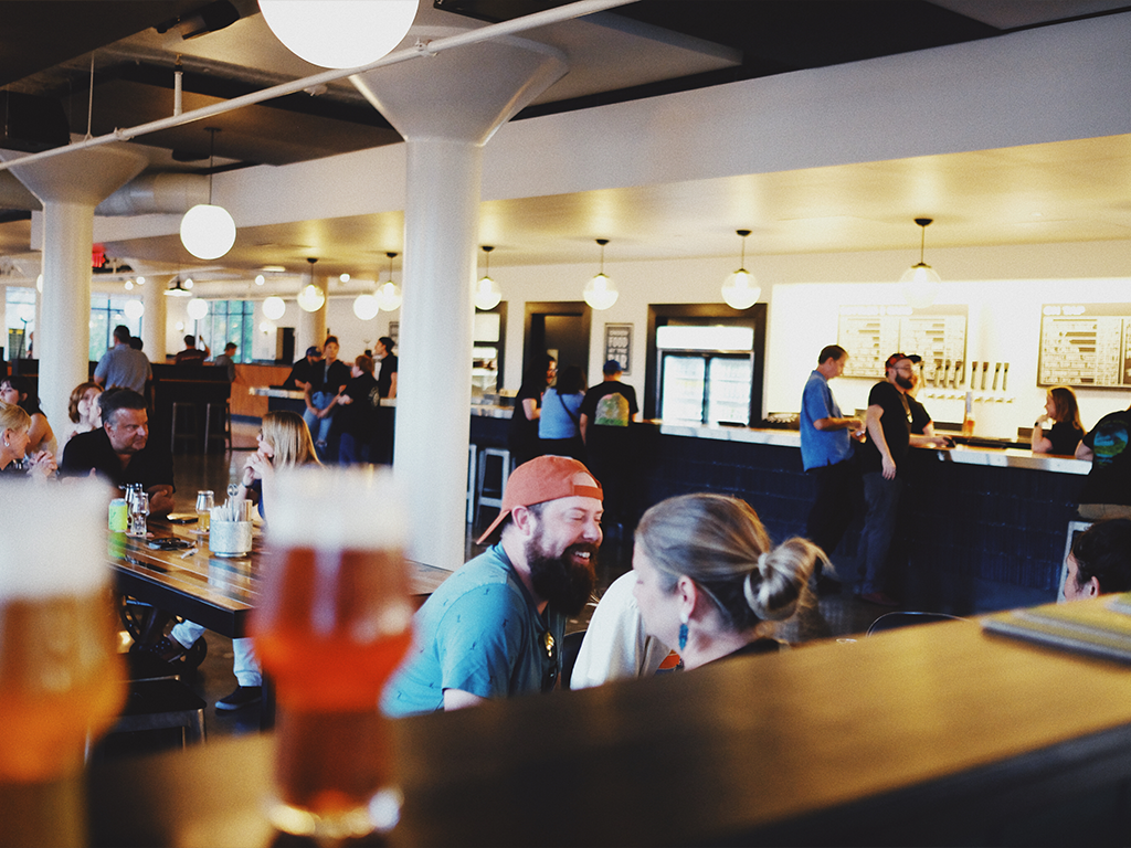 People socialize and enjoy drinks in a bright, modern bar with round ceiling lights, wooden tables, and a busy atmosphere. In the foreground, two blurred glasses of beer sit on a railing.