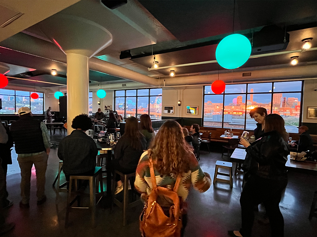 People gather in a dimly lit bar with large windows showing a cityscape at sunset. Colorful hanging lights in red, green, and blue illuminate the room, as groups socialize around tables and a musician sets up in the background.