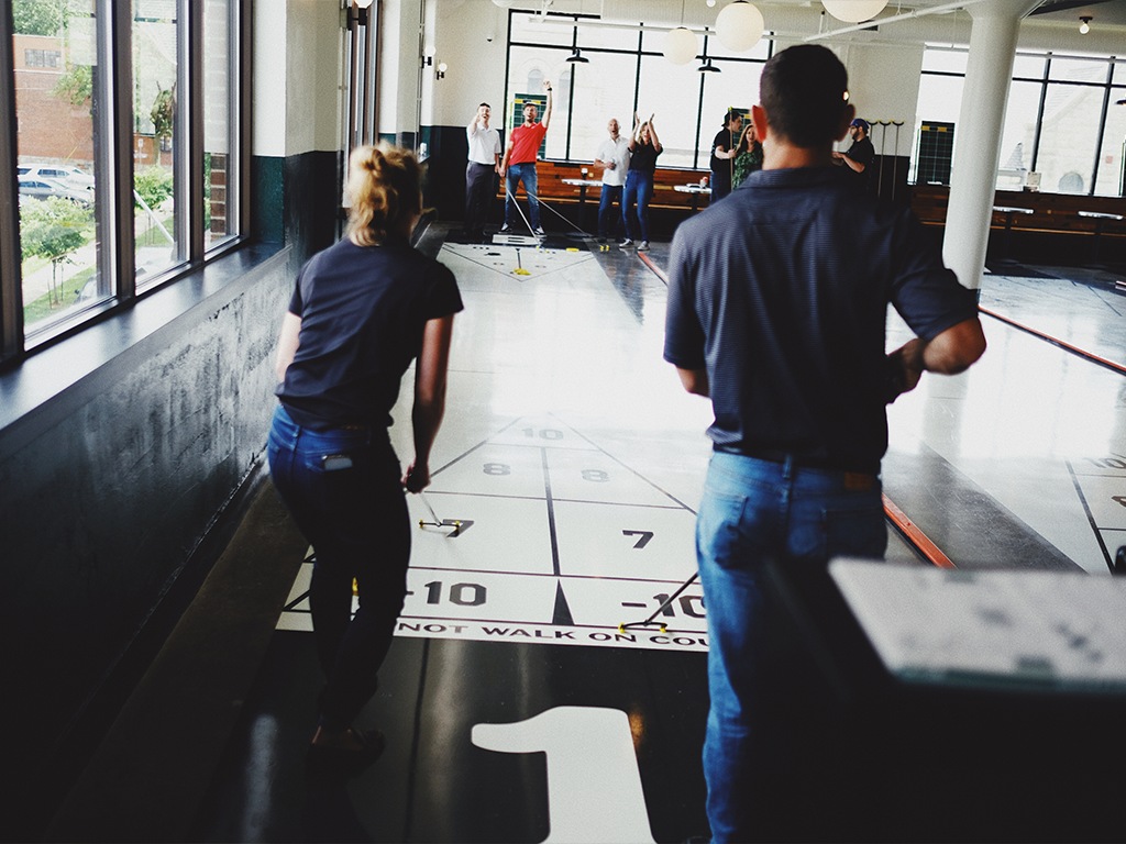 Two people in casual clothes play shuffleboard indoors, while a group of people stand and watch in the background. Sunlight streams in through large windows on the left.