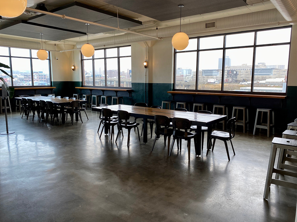 A spacious, modern room with large windows, several wooden tables and chairs, high counter seating along the windows, and round ceiling lights, offering a view of the city outside.