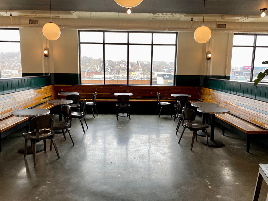 A modern, mostly empty cafe with wood and green-paneled bench seating, black chairs, round tables, and large windows letting in daylight. Spherical pendant lights hang from the ceiling.