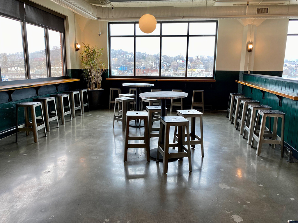 A spacious, modern room with concrete floors, metal barstools around high tables, and large windows letting in natural light. There is a potted plant in the corner and a round light hangs from the ceiling.