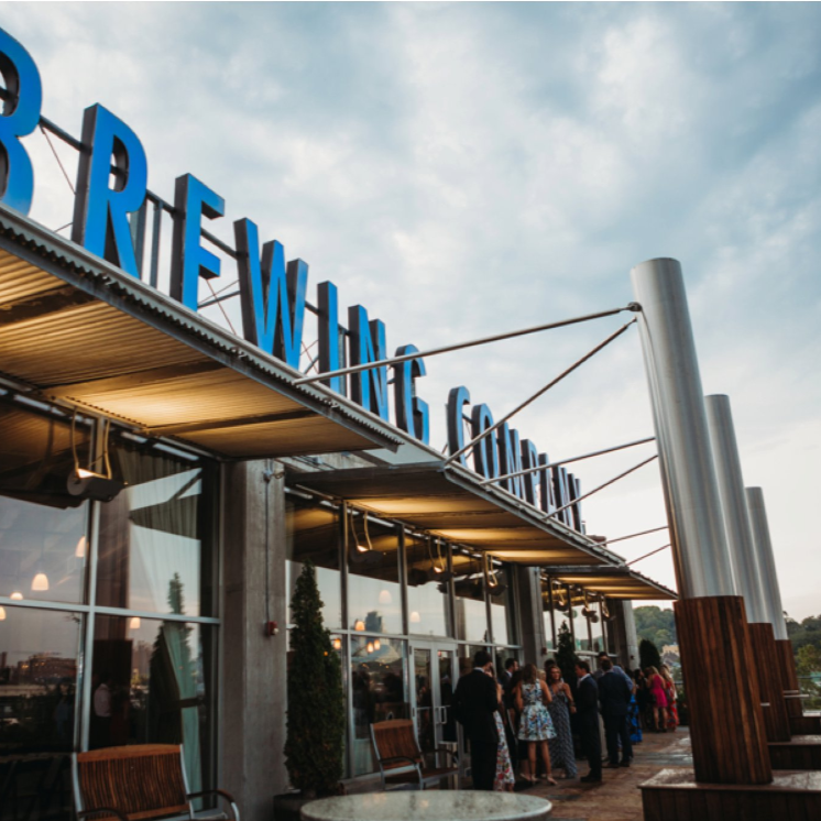 A group of people gather outside a modern building with large windows and a sign in blue letters reading "BREWING" on the roof, under a cloudy sky.