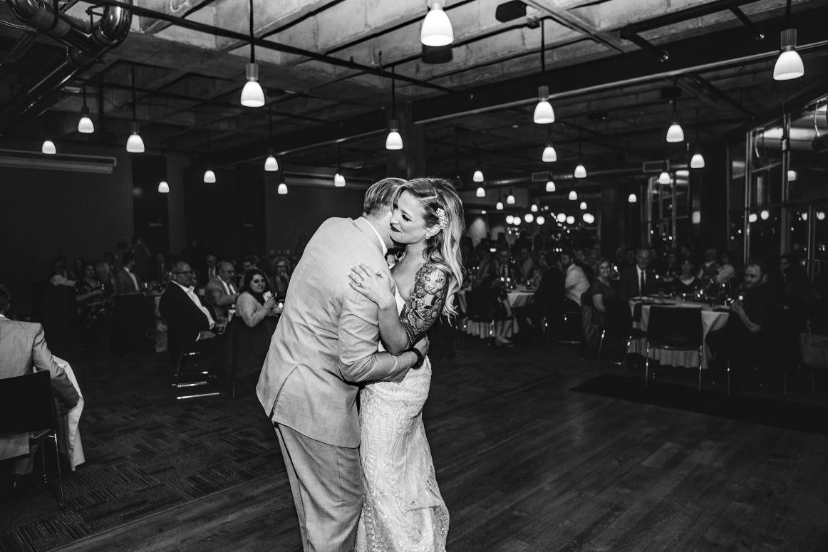 A bride and groom share a tender embrace during their first dance at a wedding reception, surrounded by seated guests in a warmly lit venue.