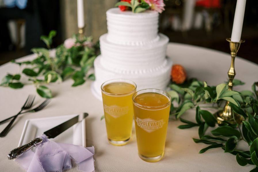 Two glasses of beer sit on a table in front of a white tiered wedding cake decorated with flowers. Greenery and a lit candle are nearby, along with utensils and a napkin on a plate.
