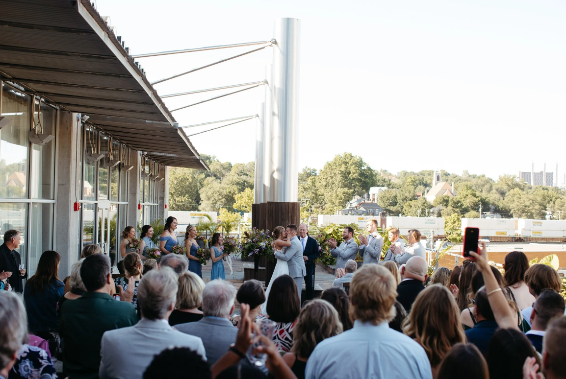 A wedding ceremony is taking place outdoors on a terrace. The couple kisses at the altar surrounded by their wedding party, while guests seated and standing in the foreground watch and take photos. Trees and buildings are visible in the background.