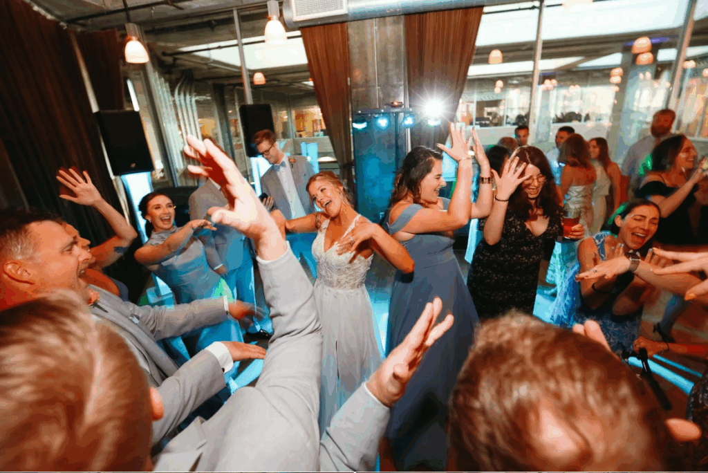 A group of people dressed in formal attire joyfully dance with raised hands at an indoor event, likely a wedding reception, under warm lighting. Everyone appears to be smiling and having fun.