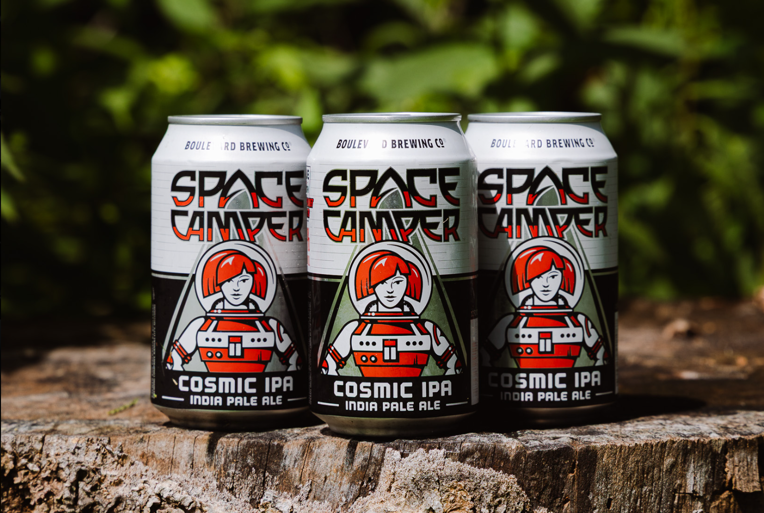 Three cans of Space Camper Cosmic IPA India Pale Ale sit on a tree stump outdoors, each adorned with the signature red-haired astronaut illustration on the labels. In the background, green foliage is artfully blurred, letting these Space Camper brews take center stage in an out-of-this-world setting.