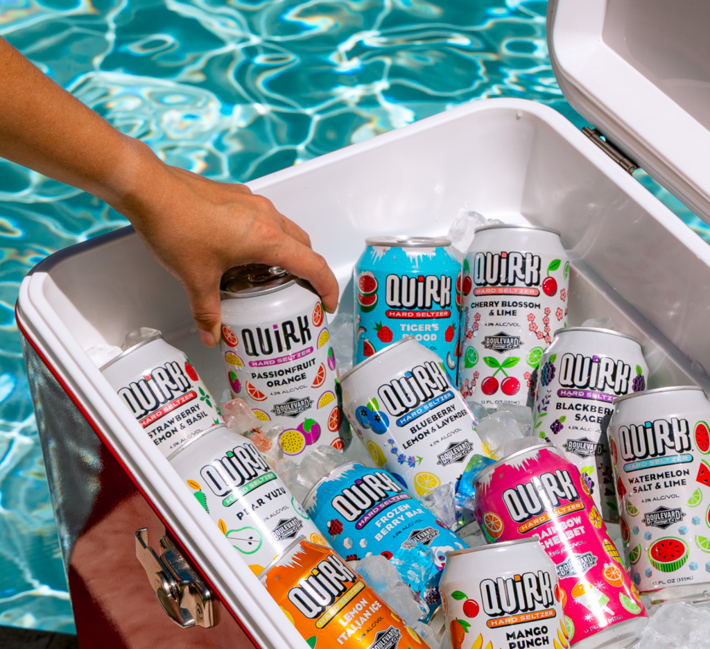 A hand reaches into a cooler filled with Quirk Hard Seltzer cans in various flavors and ice, beside a sparkling swimming pool.
