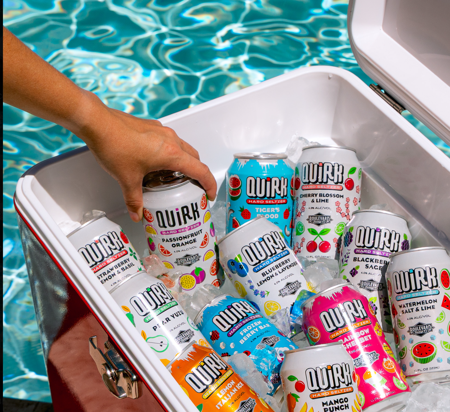 A hand reaches into a cooler filled with Quirk Hard Seltzer cans in various flavors and ice, beside a sparkling swimming pool.