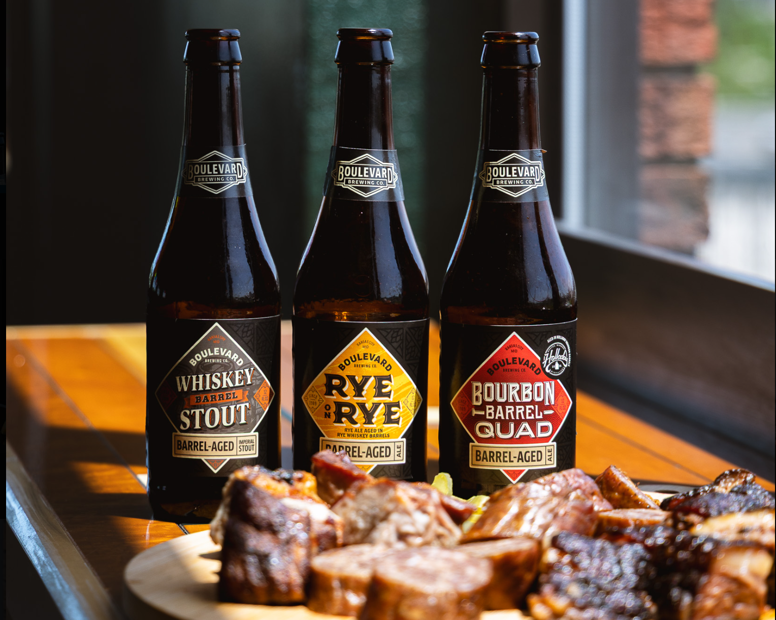 Three Boulevard Brewing Co. beer bottles—Whiskey Barrel Stout, Rye on Rye, and Bourbon Barrel Quad—stand behind a plate of assorted grilled meats on a wooden table by a window.