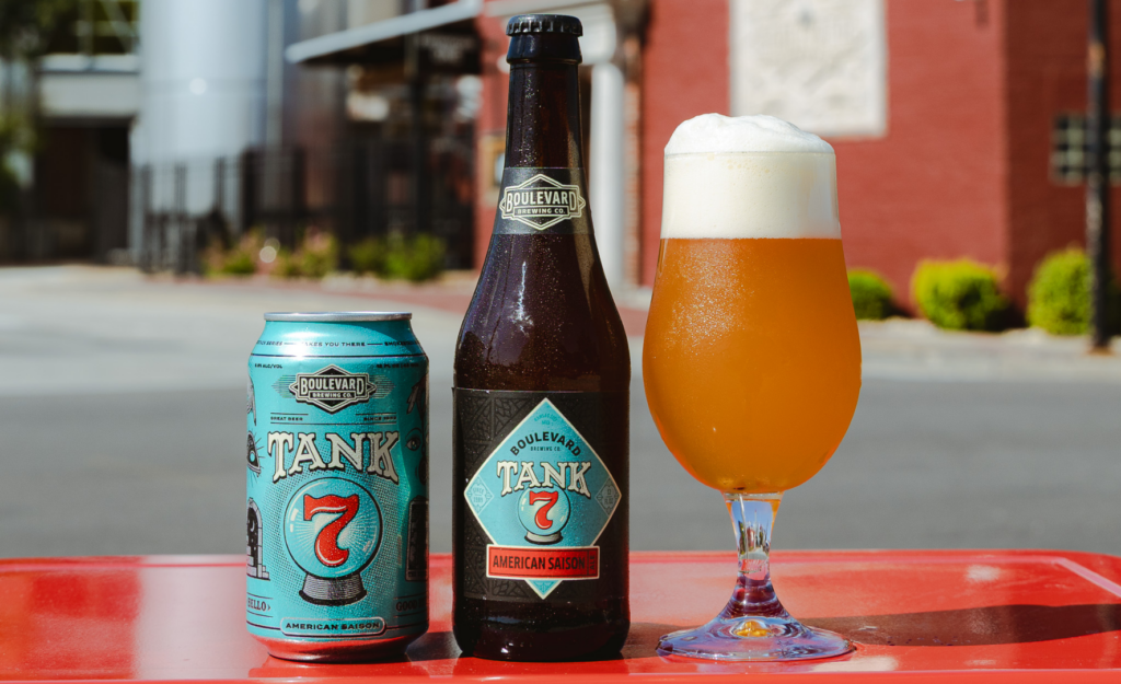 A can and bottle of Boulevard Tank 7 American Saison sit next to a glass of the beer, filled with golden liquid and a foamy head, on a red surface with a blurred outdoor background.