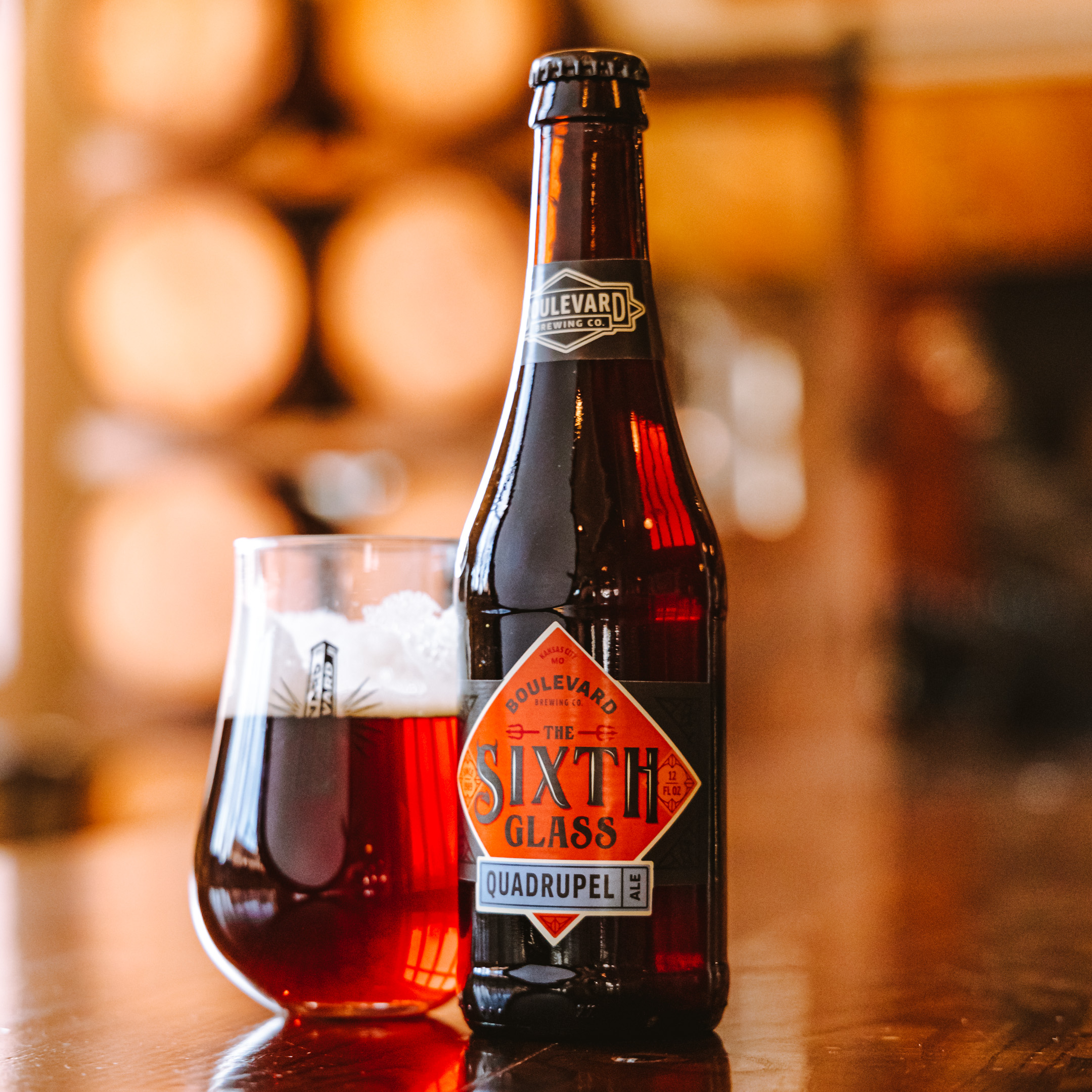 A bottle of Boulevard The Sixth Glass Quadrupel beer stands next to a glass filled with dark amber beer, set on a wooden table with blurred barrels in the background.