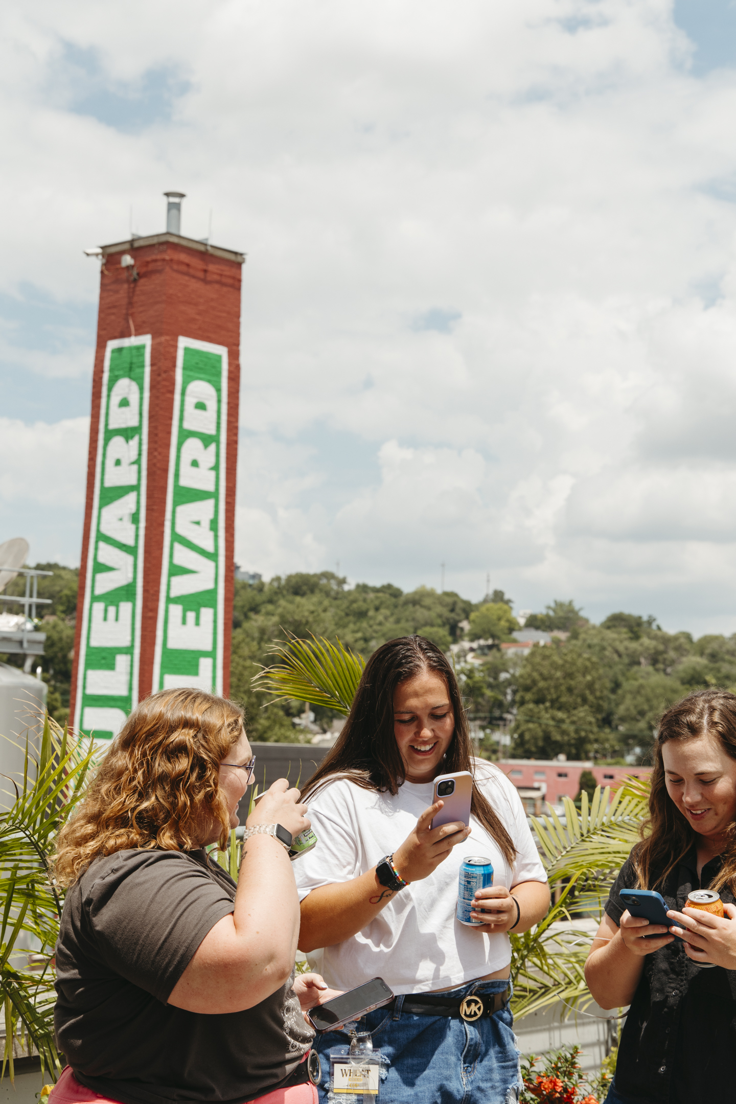 Three women stand outdoors laughing and looking at their phones, holding drinks. Behind them is a tall red brick tower with a green "Boulevard" sign—part of the Boulevard Brewing Co. Tours and Rec Center—framed by trees and a cloudy sky.