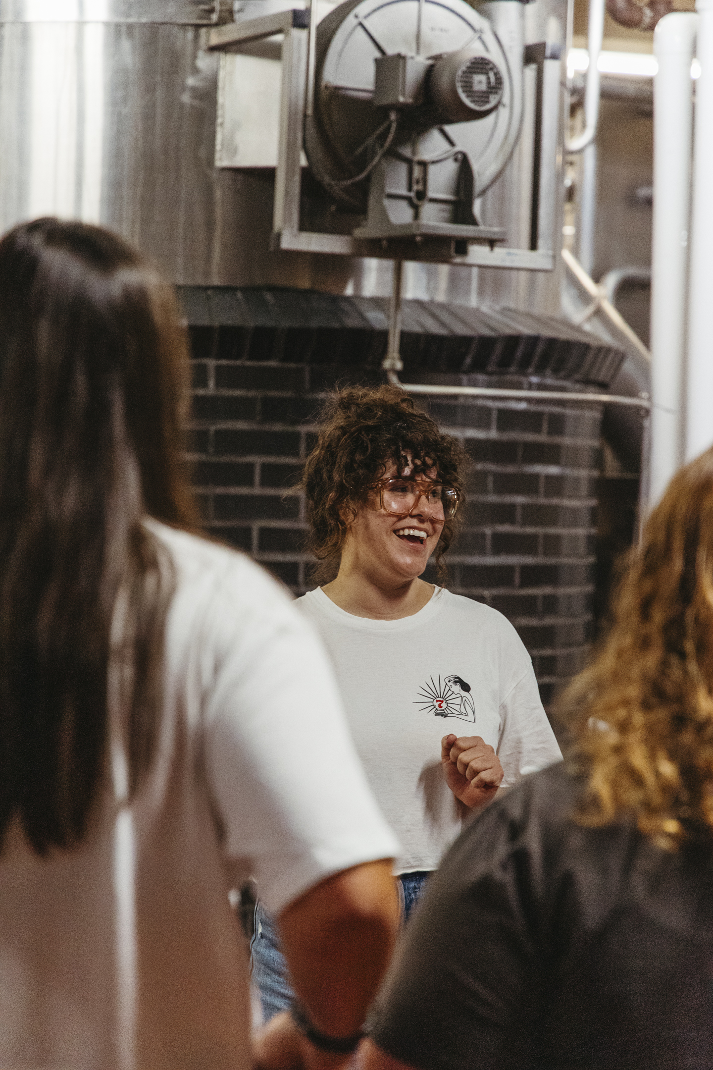 A smiling woman with curly hair and glasses in a white t-shirt stands among people on a Boulevard Brewing Co. tour in an industrial space with large metal tanks.