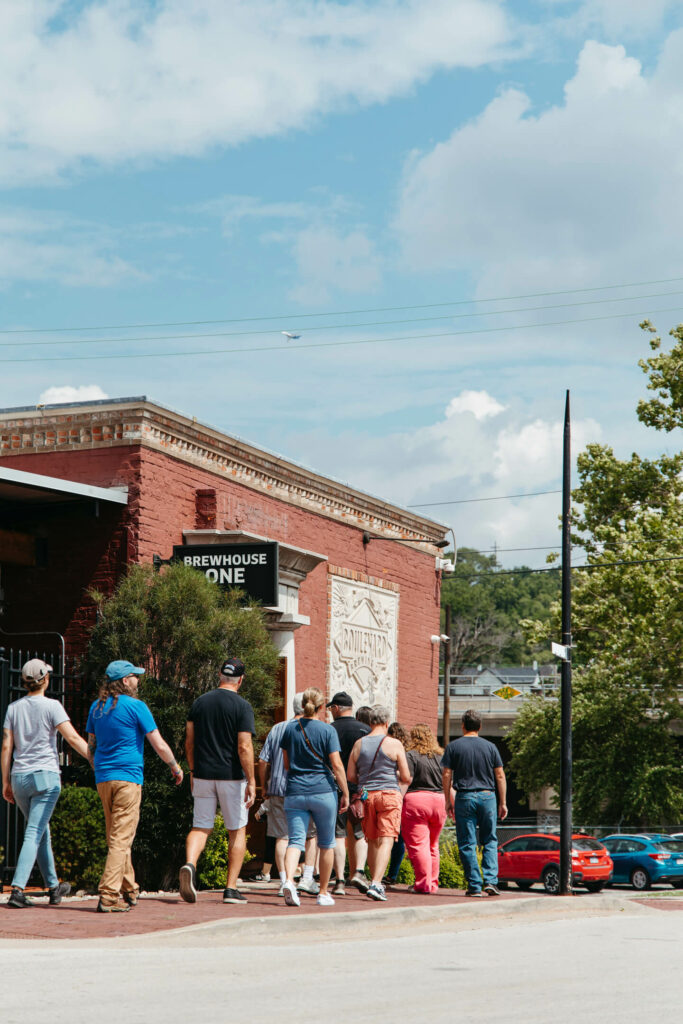 A group of people are walking towards an entrance labeled "Brewhouse One" on a red brick building. The sky is partly cloudy, and several cars are parked on the street nearby. Trees and hills can be seen in the background.