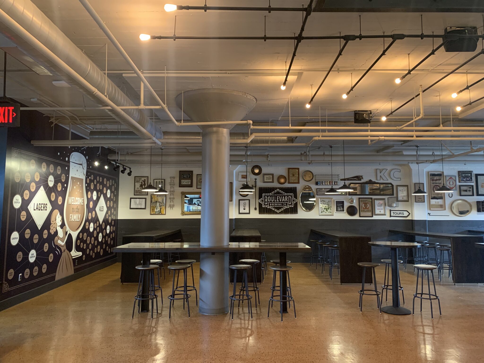 A spacious, modern taproom with high ceilings, industrial lighting, and several tall tables with stools. The walls are decorated with framed art, mirrors, and a mural featuring beer-related graphics.