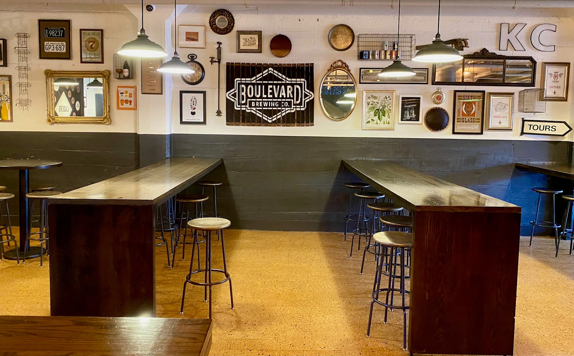 A cozy pub with long wooden tables and stools. Industrial-style pendant lights hang from the ceiling. Walls are decorated with mirrors, framed art, and a sign reading "Boulevard Brewing Co." A sign for tours is visible on the right.