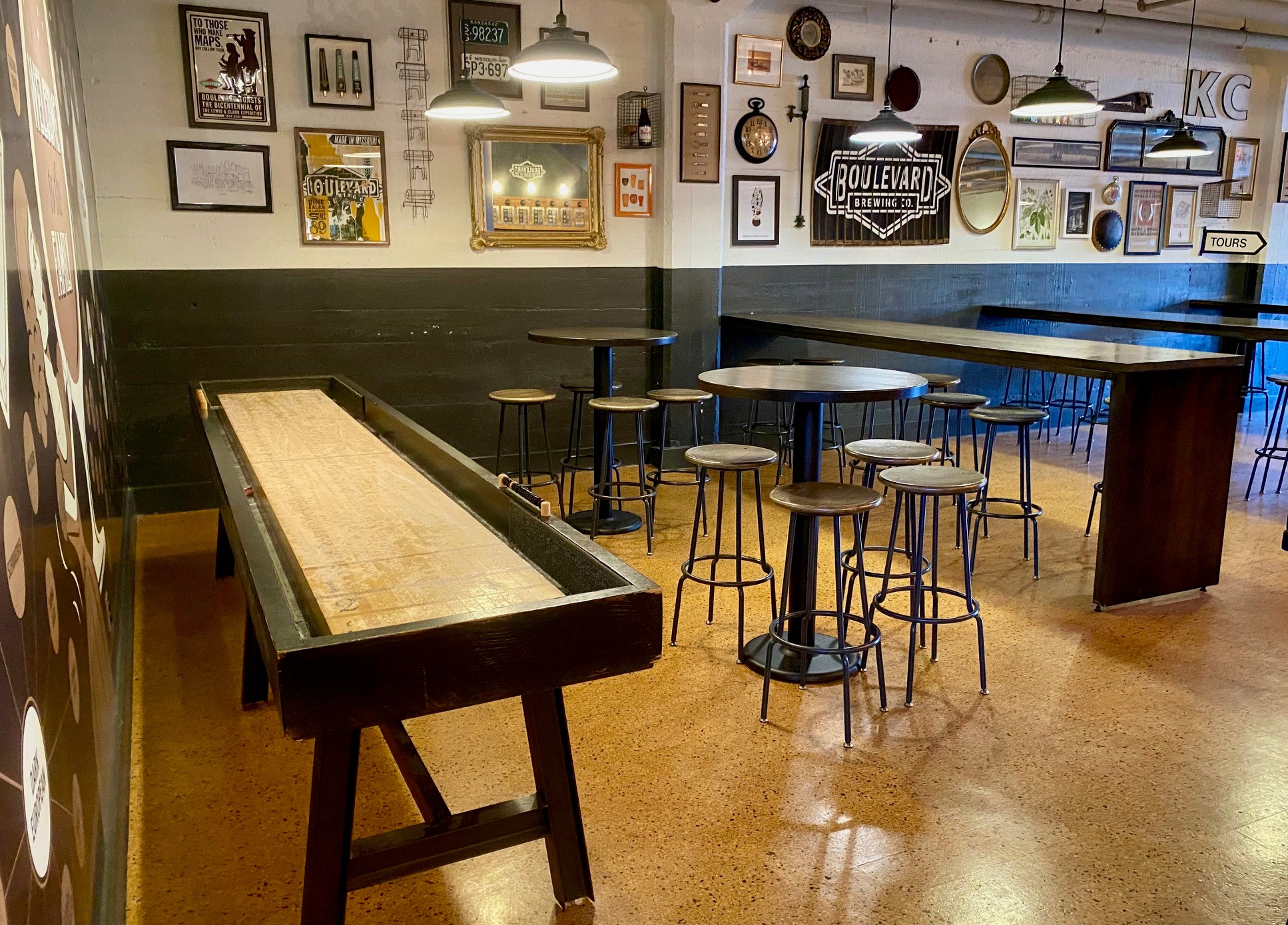 A vintage-themed interior features high tables with stools, a shuffleboard table, and various framed pictures and memorabilia on the walls. Industrial-style pendant lights hang from the ceiling, illuminating the cork flooring.