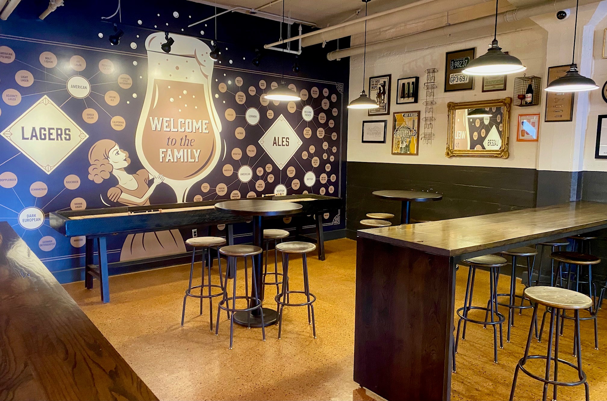 A cozy bar interior with high wooden tables and stools. The wall features a large mural with "Welcome to the Family," surrounded by labels under "Lagers" and "Ales." Framed pictures and industrial light fixtures add to the decor.
