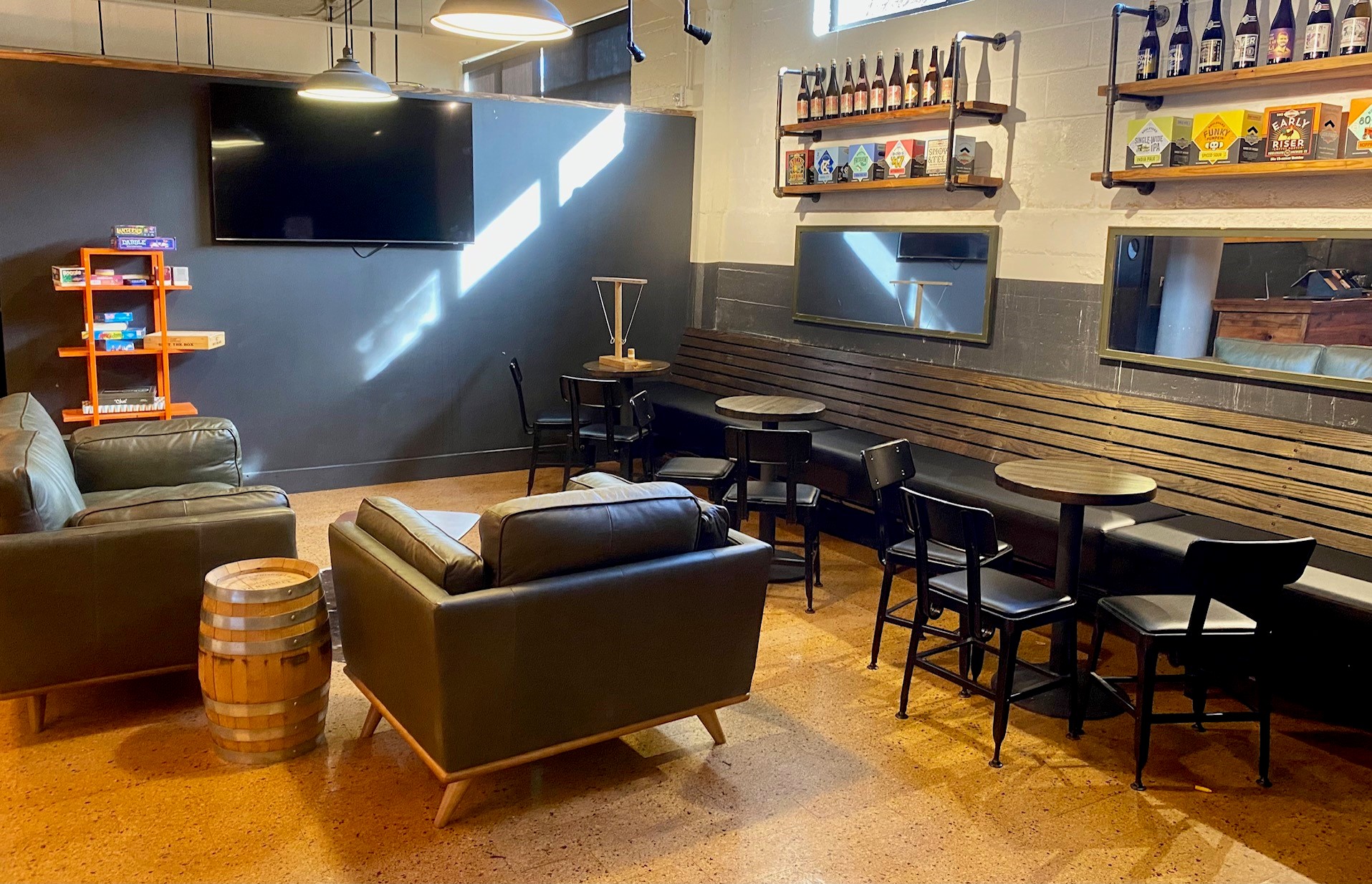 A cozy lounge area with dark leather sofas and small round tables with chairs against a gray wall. A large TV is mounted on the wall above a shelf holding board games. A wooden barrel serves as a table. Shelves display various beer bottles.