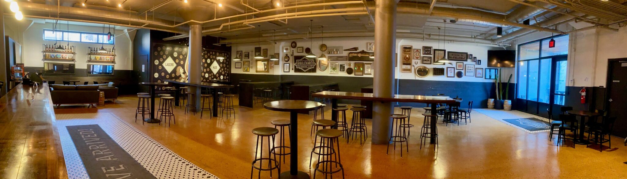 Panoramic view of a spacious, modern bar with high tables and stools, wall art and framed pictures, a bar counter on the left, and large windows letting in natural light. The space has an industrial, cozy feel.