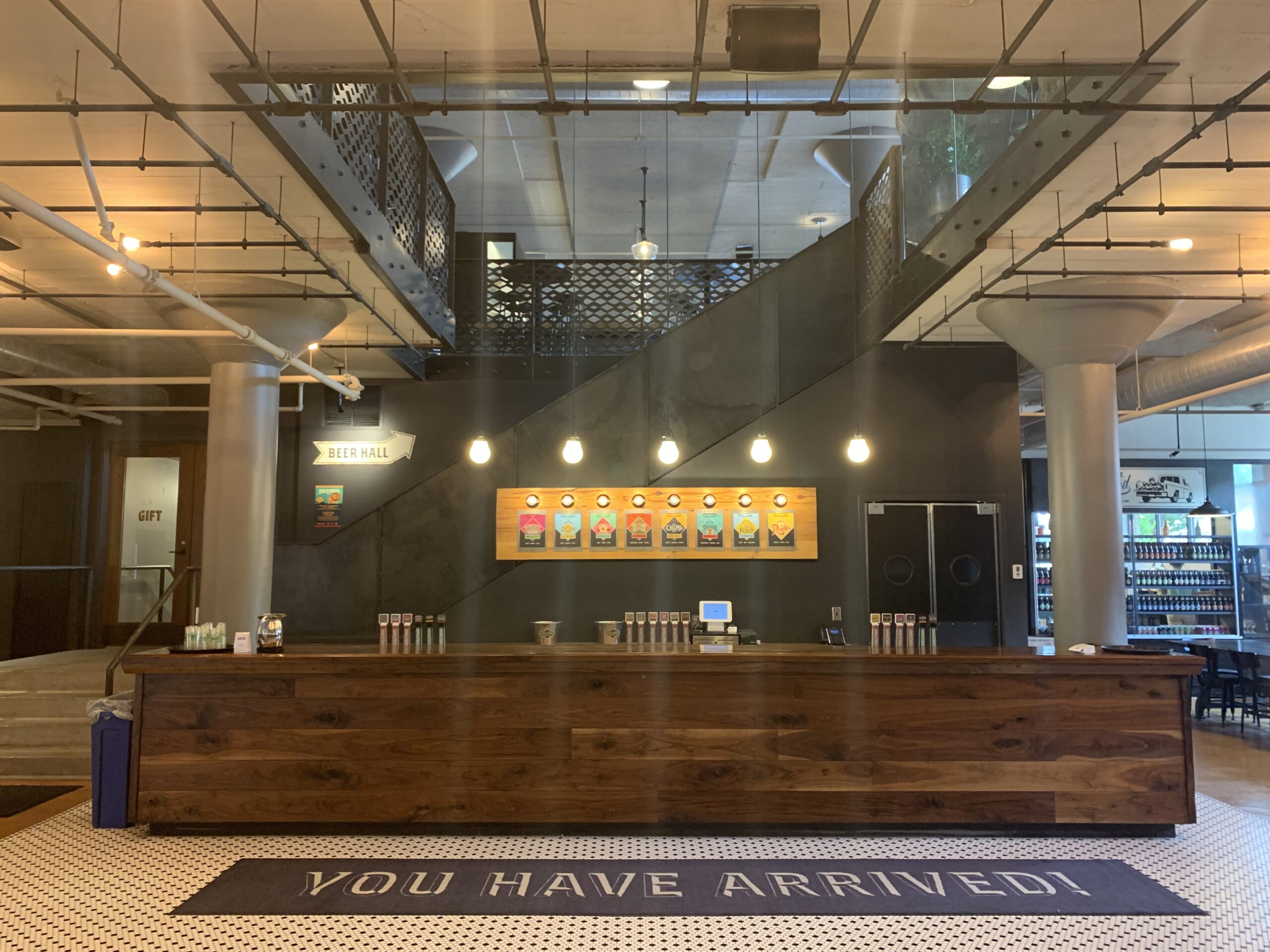 A modern brewery interior with a long wooden bar, beer taps, and a menu on the wall. Industrial décor with exposed pipes, a staircase, and a mat that reads “YOU HAVE ARRIVED!” on the tiled floor.