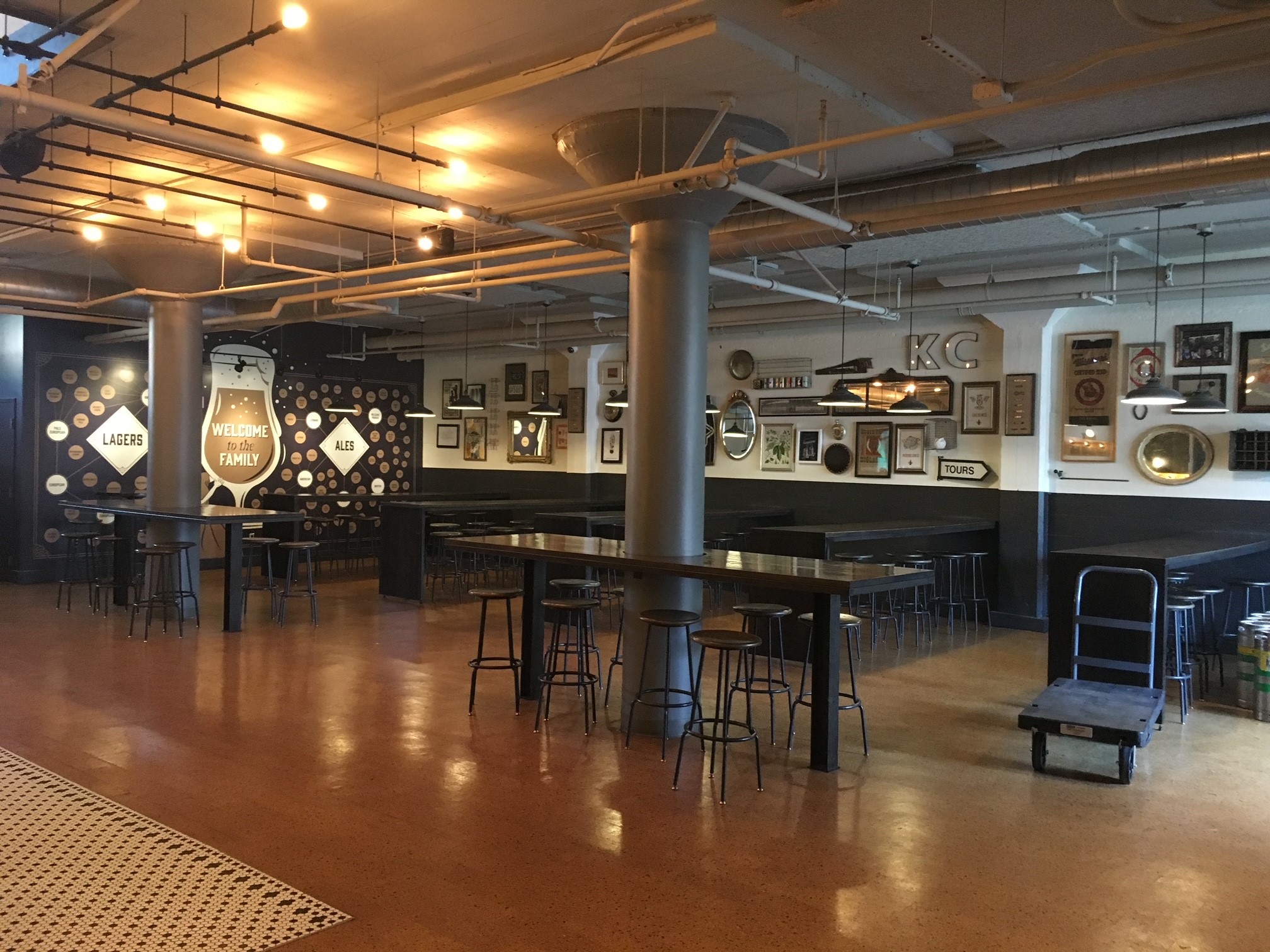 A spacious, industrial-style bar with high tables and stools, brown polished floors, exposed pipes, and walls decorated with framed pictures and signs. Warm string lights hang from the ceiling, creating a cozy atmosphere.