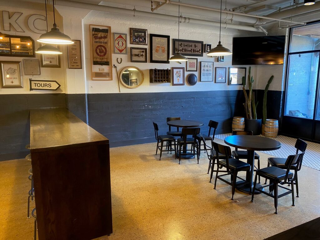A cozy lounge area with dark leather sofas and small round tables with chairs against a gray wall. A large TV is mounted on the wall above a shelf holding board games. A wooden barrel serves as a table. Shelves display various beer bottles.