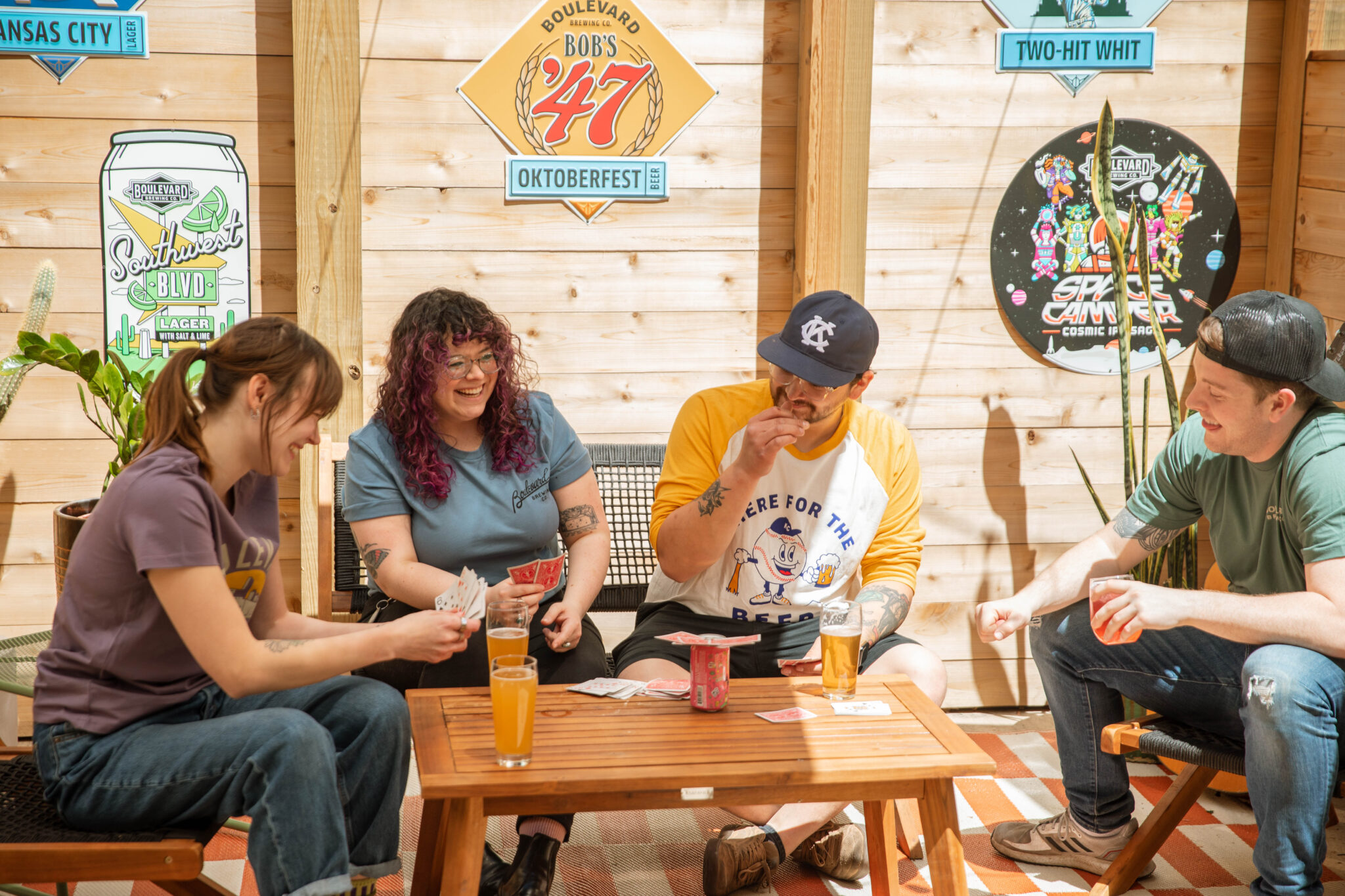 Four people sit around a small wooden table playing cards and laughing. Drinks are on the table. The setting has wooden walls decorated with colorful signs and beer-themed artwork. Sunlight brightens the cozy scene.