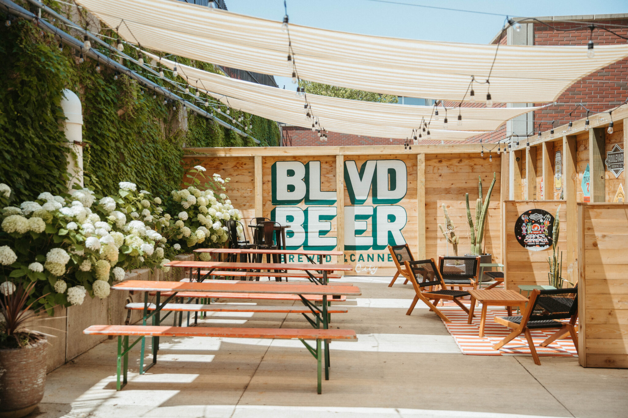 An outdoor patio with wooden picnic tables, chairs, and string lights overhead. A large “BLVD BEER” sign is displayed on a wooden wall, surrounded by greenery and blooming white flowers. The area is sunny and inviting.